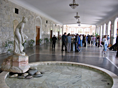 In a large hall adorned  by classical statues people drink water from fountains.