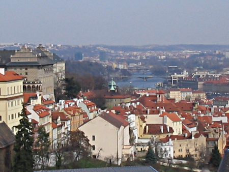 A panoramic view of Prague. There are mostly small houses, a few bredges crossing the river, and some wood among houses.