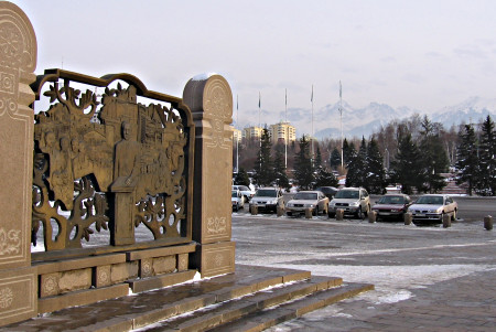 A square. On the left, a bronze bas-relief with Presidenz Nazarbaev swearing on the constitution. Snowy mountain tops in the background.