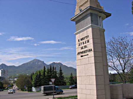 A large stone pillar with "To the place of Lermontov's duel" written on it in Russian. In the backround, streets and mountains.