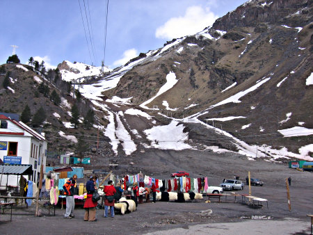 There are a few cars and a few sellers of traditional furs and clothing. Ski lift wires go up the partially snowed slope of the mountain.