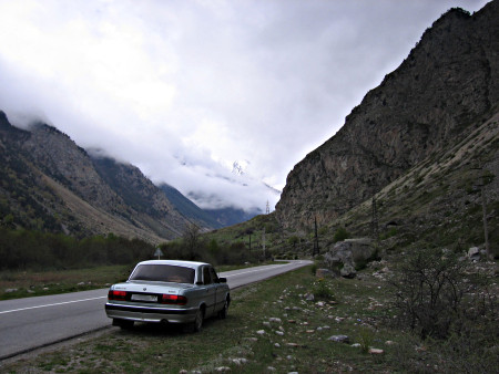 A large gray Volga car parked next to a road. The road passes through a rocky valley, the sky is very cloudy.