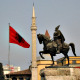 Albanian flag is waving next to a statue of a warlord riding a horse, and in front of a minaret.
