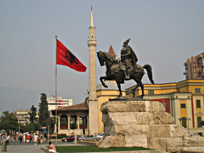 Albanian flag is waving next to a statue of a warlord riding a horse, and in front of a minaret
