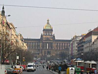A large boulevard with cars passing on both sides and a pedstrian area in the middle constitute the square. In the background, the large building of the National Museum.
