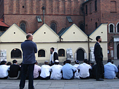 A group of young people wearing the traditional Jewish headcloth sit next to a synagogue and listen to a rabbi.
