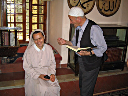 A smiling nun in a long and ligth gown is discussing with a man wearing a muslim cap and holding a book in his hands
